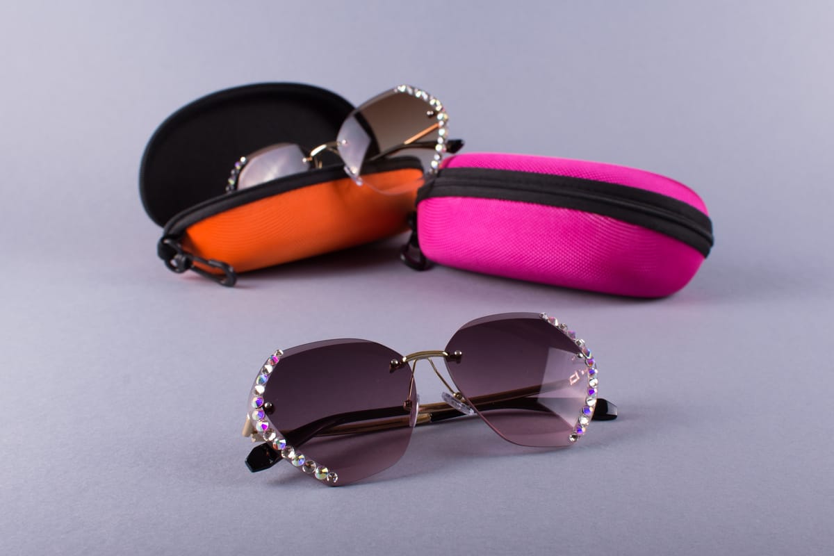 Occhiali con lenti viola sfumate in primo piano e due custodie rigide, arancione e fucsia, sullo sfondo