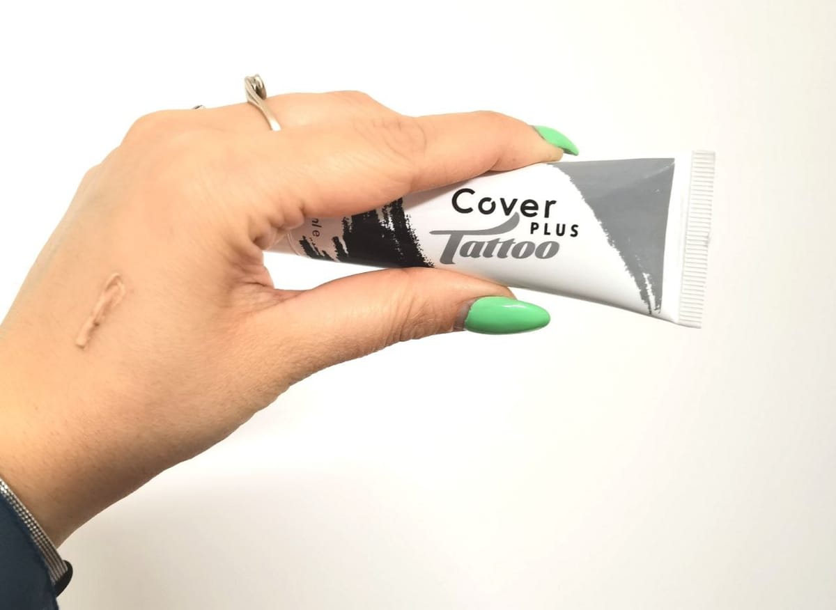 Una mano tiene il tubetto CoverPlus Tattoo mentre sul dorso è stesa una striscia di crema color incarnato